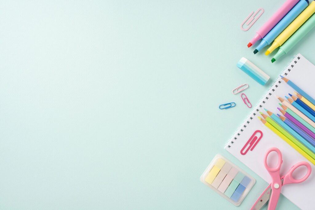 Flat lay of colorful school stationery including scissors, pencils, markers, paper clips, and a notebook on a light blue background