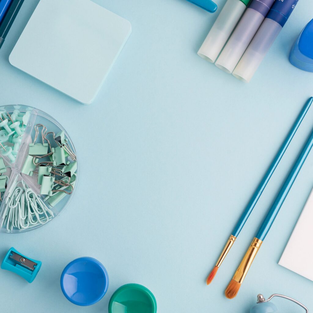 Organized flat lay composition featuring blue and green school stationery items, including pens, markers, scissors, sticky notes, notebook, paintbrushes, clips, and a small alarm clock arouged as round